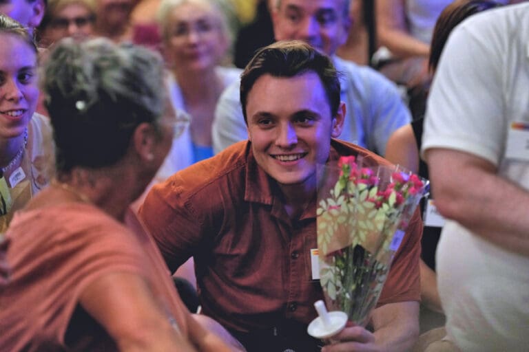 A man beams with happiness as he holds flowers, engaging with a lively crowd surrounding him.